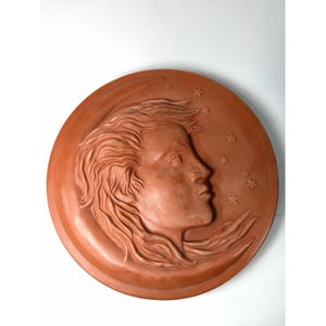 Pode incluir: Uma placa de parede circular de terracota com uma escultura em relevo de um rosto de perfil com cabelo fluido, integrado numa forma de meia-lua. O design inclui várias pequenas estrelas de cinco pontas na superfície lisa.
