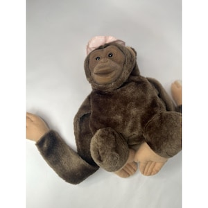 May include: A plush orangutan toy with dark brown fur, a light tan face, hands, and feet, and a small, light pink fabric accessory resting on the top of its head. The stuffed animal features long arms and a soft, textured body design.