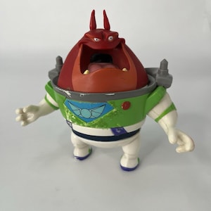 Op de afbeelding: Een plastic actiefiguur van een rood, oranje, groen en wit monster met een open mond en kleine hoorns. De figuur draagt een groen en wit ruimtepak met een blauw en wit embleem. De figuur is een personage uit de Toy Story-franchise.