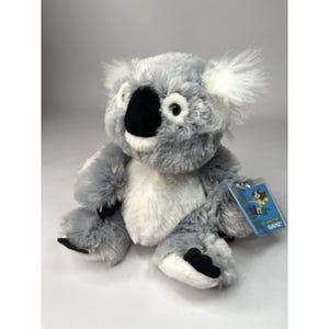 May include: A plush koala bear with gray and white fur. The koala has a black nose, black paws, and a tag attached to its ear. The tag has the words "Webkinz" and "GANZ" printed on it.