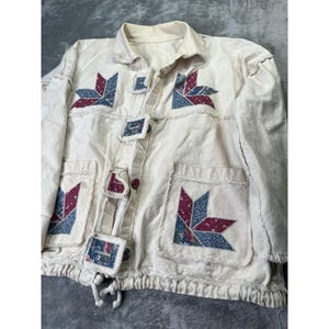 May include: Cream-colored jacket with patchwork star designs in red and blue. Features square button closures and two front pockets. The jacket has a drawstring hem and a collar.