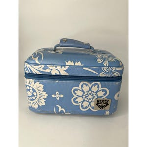May include: A blue cosmetic bag with a white floral pattern and a top handle. The bag has a zipper closure and a Roxy logo on the front. The bag is approximately 8 inches wide, 6 inches tall, and 5 inches deep.