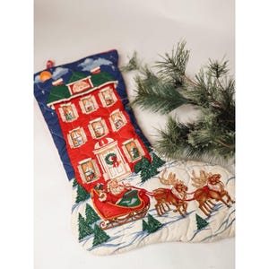 May include: A quilted Christmas stocking featuring a dark blue night sky background with a tall red house, a scene of Santa Claus in a red sleigh pulled by two reindeer, and green pine trees on a white snowy base with a pine branch accent.