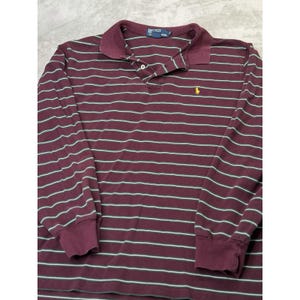 May include: A long-sleeve polo shirt in a deep burgundy color featuring thin horizontal stripes in white and light blue. The shirt has a collar, a buttoned placket, and a small yellow embroidered logo on the left chest. A blue label is at the neck.