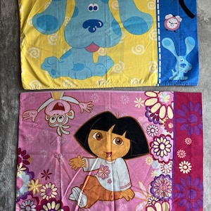 Puede incluir: Dos fundas de almohada rectangulares con personajes de dibujos animados. Una presenta un perro azul sobre un fondo amarillo. La otra tiene a Dora la Exploradora y un mono sobre un fondo rosa. También hay patrones florales y otros elementos de dibujos animados.