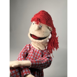 May include: A hand puppet featuring a beige fabric face with freckles, googly eyes, and a red yarn wig styled in a side ponytail. The puppet wears a long-sleeved shirt made of red, blue, and white plaid fabric with a round neckline.
