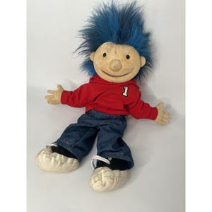 Puede incluir: Una muñeca de peluche con una cara bronceada, cabello azul y una sudadera con capucha roja con el número 1. La muñeca lleva vaqueros azules y zapatillas blancas con detalles en negro. Los brazos de la muñeca están extendidos.