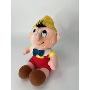 Può includere: Bambola di peluche di Pinocchio, con cappello giallo, capelli neri e naso lungo. La bambola indossa un colletto bianco, un papillon blu, una camicia gialla, pantaloncini rossi e scarpe marroni. La bambola è seduta.