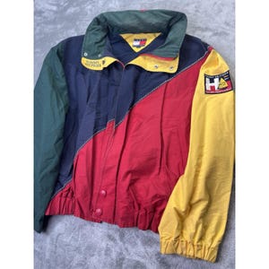 May include: A color-blocked windbreaker jacket featuring navy blue, red, yellow, and dark green panels. The jacket includes a high collar, a front zipper closure, and a Tommy Hilfiger Sailing Gear patch on the right sleeve with the text H and TH.