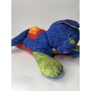 May include: A stuffed cat toy crafted from patchwork fabric featuring a primary blue base with swirling patterns. The design incorporates contrasting segments in lime green, bright orange, and red, with embroidered yellow eyes and a purple nose.