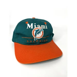 May include: A teal and orange baseball cap with the words "Miami Dolphins". The cap has a curved peak and a logo of a dolphin leaping through a circle. The cap is made of a durable fabric.