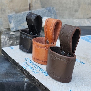 May include: Three leather knife sheaths in black, tan, and brown. Each sheath has a loop for a belt and decorative embossed patterns around the top and bottom. The sheaths are made of thick leather and have a metal rivet.
