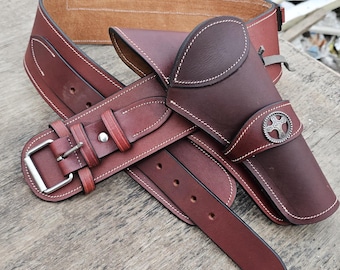Könnte beinhalten: Ein braunes Leder-Gürtel- und Holster-Set. Der Gürtel hat eine silberne Schnalle und weiße Nähte. Das Holster hat ein dekoratives silbernes Emblem und ist für eine Schusswaffe konzipiert. Das Set ist aus hochwertigem Leder gefertigt.