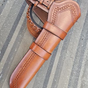 May include: A brown leather gun holster with a matching belt. The holster features intricate embossed patterns and secure straps. The belt has a silver buckle. The set is designed for carrying a firearm and is likely for a Western-themed outfit.