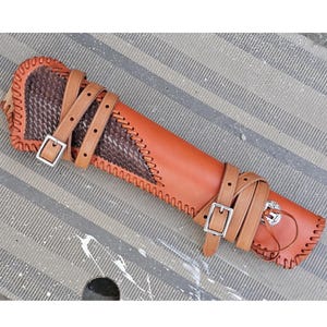 May include: A handcrafted leather quiver in shades of brown. The quiver features a woven leather accent, decorative stitching, and adjustable straps with silver buckles. The design includes a decorative metal emblem and is suitable for archery.