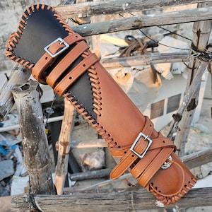 May include: A brown leather archery arm guard with a black leather backing. The guard features decorative stitching, adjustable straps with silver buckles, and a rounded bottom. The design is rustic and functional.