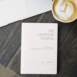 Op de afbeelding: Een crèmekleurig tijdschrift met de titel "The Gratitude Journal" met de tekst "A 30-Day Guide to Awareness, Joy, and Inner Growth." Een laptop en een kopje koffie met latte art staan ook op het donkere houten oppervlak.