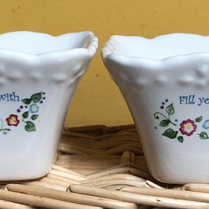 Puede incluir: Dos macetas cuadradas de cerámica blanca con bordes festoneados y diseños florales. Una dice "Fill your Home with Happiness", la otra "Fill your Heart with Love". Están sobre una cesta tejida, ideal para plantas pequeñas.