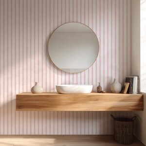 May include: A modern bathroom with a round mirror, a white sink, and a wooden shelf. The wall features a pink and white striped wallpaper. Decorative vases, books, and a woven basket add to the decor.