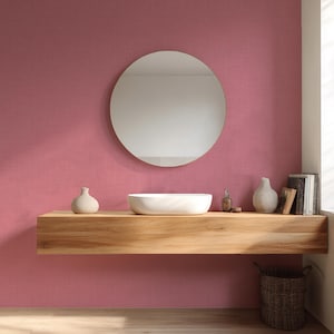 May include: A modern bathroom scene featuring a round mirror with a gold frame, a floating wooden shelf, and a white vessel sink. The shelf holds decorative vases, a soap dispenser, and books. The walls are painted a dusty rose color, and a woven basket sits below.