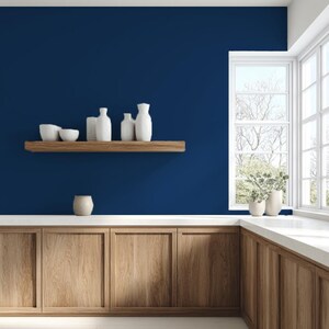 May include: A modern kitchen with a deep blue wall and wooden cabinets. A wooden shelf displays white vases and bowls. A small, beige ceramic vase sits on the countertop. A window with white trim and a view of trees is in the background.