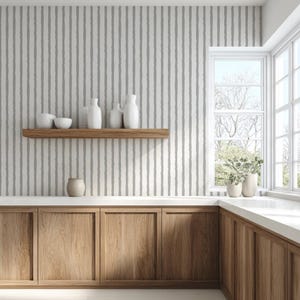 May include: A kitchen interior with a light gray and white striped wallpaper. A wooden shelf holds white vases and bowls. Below, wooden cabinets with a white countertop and a window with natural light complete the scene.