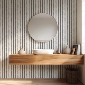 May include: A modern bathroom scene featuring a round mirror with a gold frame above a wooden floating shelf. The shelf holds a white vessel sink, decorative vases, and a soap dispenser. The wall is covered in vertical gray and white striped wallpaper.