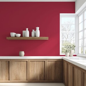 May include: A kitchen interior with a deep red wall, wooden cabinets, and a white countertop. A wooden shelf displays white vases and bowls. A window offers a view of trees and greenery. A small, beige ceramic vase sits on the countertop.