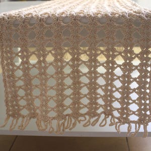 Può includere: Runner da tavolo all'uncinetto beige, a trama aperta, con motivo geometrico e dettagli a frange. Il runner è drappeggiato su un tavolo bianco, mettendo in risalto il suo design e la sua trama complessi. Questo pezzo decorativo aggiunge un tocco di fascino fatto a mano.