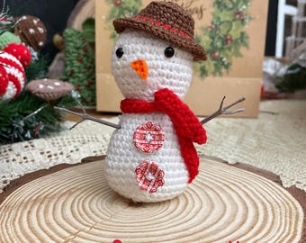 Peut inclure: Un bonhomme de neige crocheté avec un chapeau marron, une écharpe rouge et des bras en brindilles. Le bonhomme de neige a un nez en carotte et est décoré de boutons rouges et blancs. Le texte "Snowman pattern" est visible.