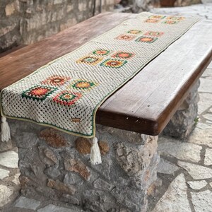 May include: Cream crochet table runner featuring colourful square patterns in orange, green, and yellow. The runner has white tassel fringe at each end. The table runner is displayed on a wooden table.