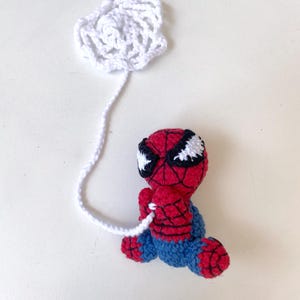 May include: A handmade crochet Spider-Man doll in a sitting position, with a white web and string. The doll features a red and black mask, red body, and blue pants. The web is white and attached to the doll.