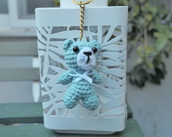 May include: A handmade, light blue crochet teddy bear keychain with a white muzzle and a small white bow. The bear has black eyes and a gold-colored chain. The keychain is hanging in front of a white, decorative lantern.