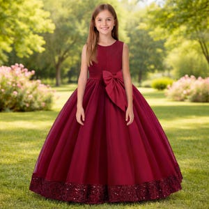 May include: A floor-length sleeveless dress in deep burgundy featuring a fitted bodice with vertical pleats, a large bow at the waist, and a full skirt with a wide band of matching sequins along the hemline, constructed from satin and tulle fabric.