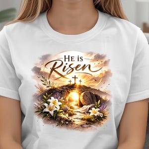May include: White t-shirt with the words "He is Risen" in a decorative font. The design shows a sunrise, three crosses, and a tomb, with floral elements. The artwork has a watercolour style.