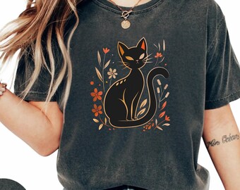 Può includere: T-shirt grigio scuro con un'illustrazione di un gatto nero circondato da fiori e foglie arancioni e bianche. Il gatto ha gli occhi gialli ed è seduto. La parola "PEPPER" è stampata su un cerchio grigio scuro in basso.
