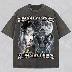 May include: Dark gray t-shirt featuring a graphic of three werewolves with a full moon backdrop. The text "HUMAN BY CHANCE" and "ALPHA BY CHOICE" are printed across the top and bottom of the design. The shirt has a vintage, distressed look.