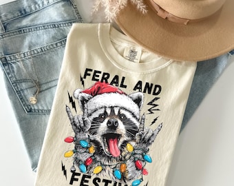 Può includere: T-shirt color crema con grafica di un procione con cappello di Babbo Natale e luci natalizie, con la scritta "FERAL AND FESTIVE". La maglietta è appoggiata su jeans azzurri e un cappello beige.