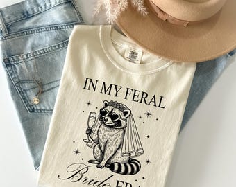 May include: Cream-colored t-shirt with the text "IN MY FERAL Bride ERA" and a raccoon graphic wearing a veil and heart-shaped sunglasses. The shirt is laid on top of light blue jeans, a gold necklace, and a tan hat.