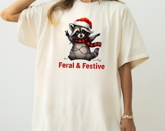 Può includere: T-shirt color crema con un procione che indossa un cappello di Babbo Natale e una sciarpa a quadri rossi e neri. Il procione è decorato con luci natalizie e la scritta "Feral & Festive" in rosso.