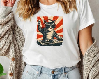 May include: White t-shirt featuring a cartoon cat DJ wearing sunglasses and headphones, with a red and cream-colored background. The cat is positioned behind a turntable, with one paw raised.