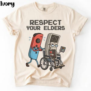 May include: Ivory t-shirt featuring the text "RESPECT YOUR ELDERS" in a pixelated font. The graphic depicts a red and blue game controller pushing a gray controller in a wheelchair. The gray controller holds a game cartridge.