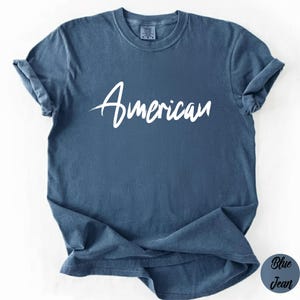 May include: Blue jean-colored t-shirt with the word "American" in white script across the front. The shirt has rolled-up sleeves and a relaxed fit. The tag on the back of the neck is visible.
