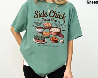 May include: Light green t-shirt with the text "Side Chick Social Club" and a graphic of various side dishes. The design includes a gravy boat, rolls, mashed potatoes, fried food, green beans, cranberries, and small cups. The phrase "I'M JUST HERE FOR THE SIDES" is printed below.
