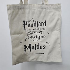 Peut inclure: Un sac fourre-tout en toile beige avec du texte noir qui dit "Poudlard ne recrutait plus DU COUP j'enseigne aux Moldus." Le design comprend un château, des éclairs et une baguette. Le sac a deux poignées.