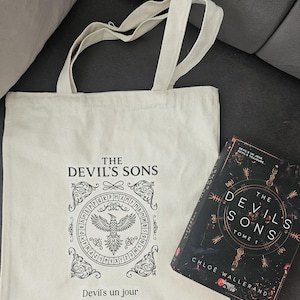 Peut inclure: Un sac fourre-tout crème et un livre intitulé "The Devil's Sons" sont présentés. Le sac fourre-tout présente un graphisme noir avec le texte "The Devil's Sons" et "Devil's un jour Devil's toujours". Le livre a une couverture sombre avec le même titre.