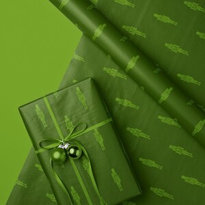 Op de afbeelding: Groen cadeaupapier met een herhalend patroon van notenkrakerfiguren. Een ingepakt cadeau is versierd met een groen lint en twee groene ballen. Een rol bijpassend papier is zichtbaar, alles op een groene ondergrond.