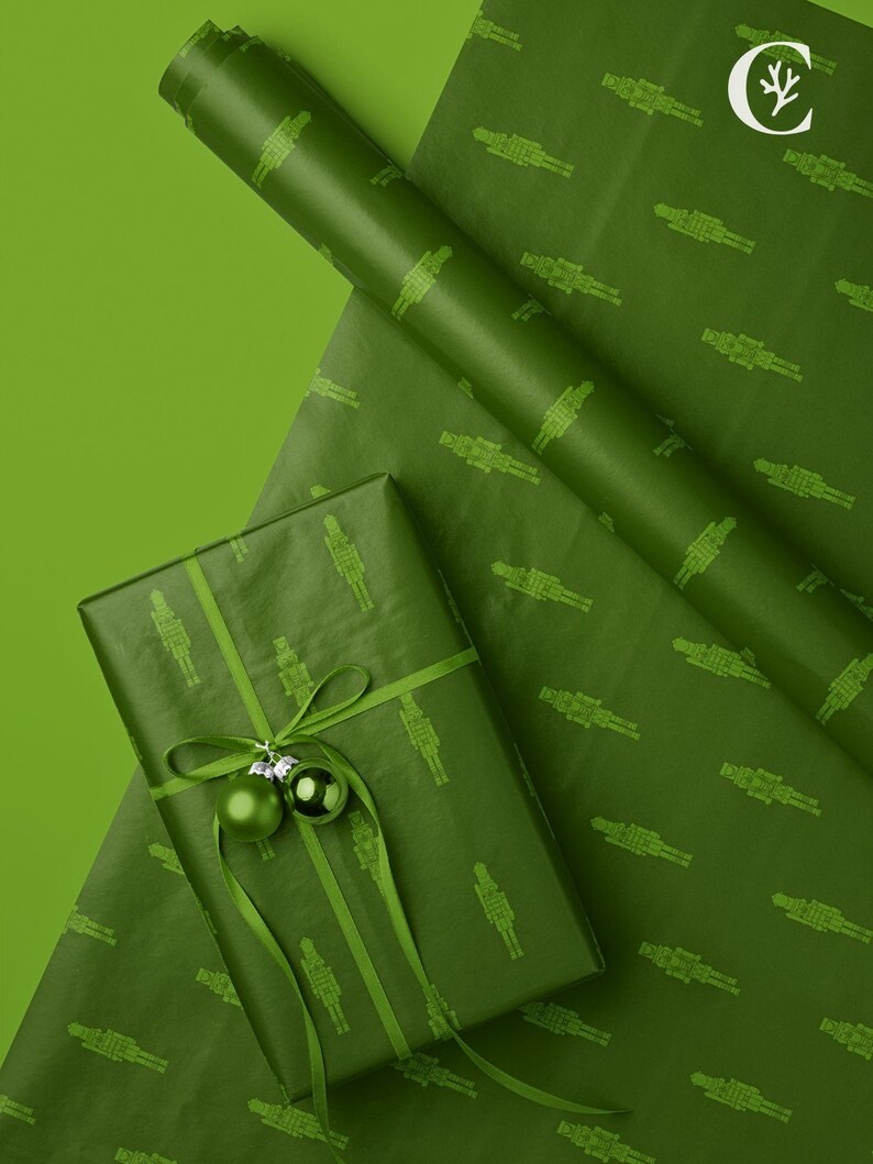 Op de afbeelding: Groen cadeaupapier met een herhalend patroon van notenkrakerfiguren. Een ingepakt cadeau is versierd met een groen lint en twee groene ballen. Een rol bijpassend papier is zichtbaar, alles op een groene ondergrond.