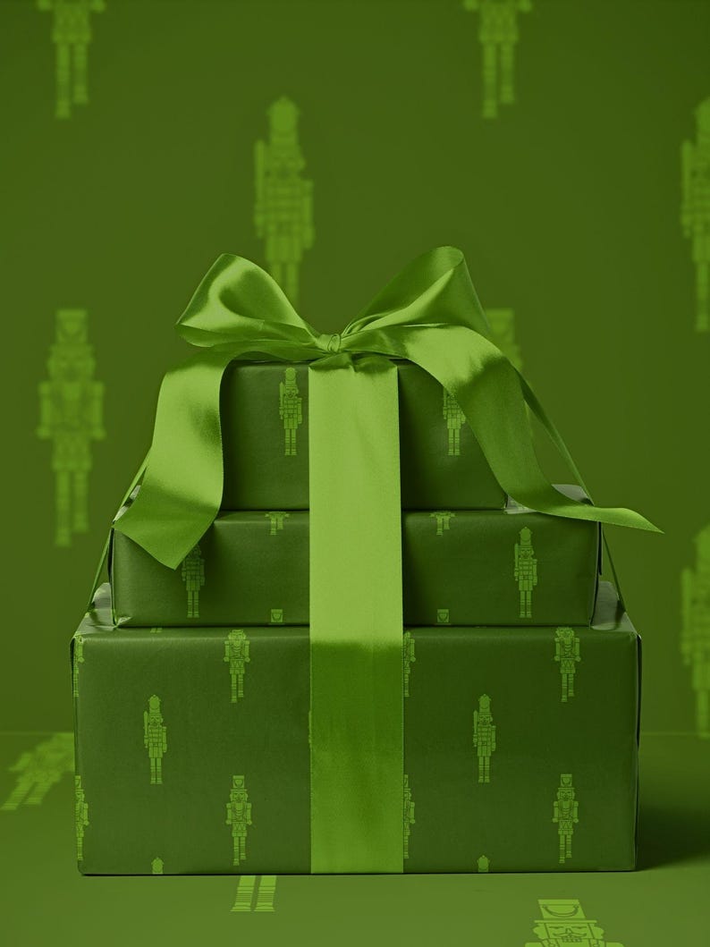 Op de afbeelding: Drie gestapelde, verpakte cadeaus in groentinten, vastgebonden met een bijpassend groen satijnen lint en strik. Het cadeaupapier heeft een herhalend patroon van silhouetten van notenkrakersoldaten. De achtergrond is effen groen.