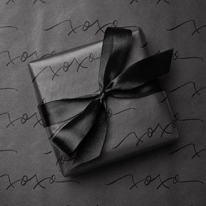 May include: A wrapped gift with a black satin ribbon bow. The gift is wrapped in black paper with a repeating pattern of the word "xoxo" in a handwritten style. The overall aesthetic is minimalist and elegant.